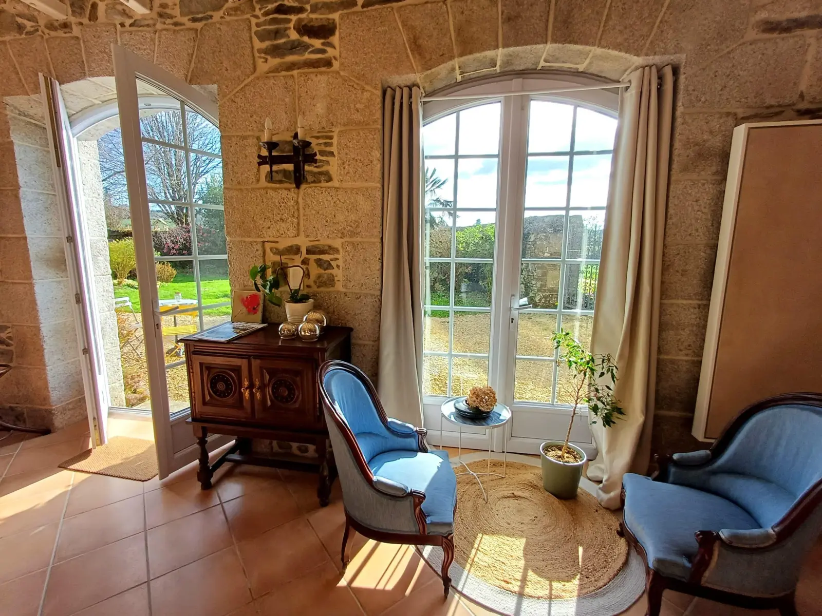 Sitting room with stone walls, blue armchairs, wooden chest and French windows open to the garden, Ty Monde