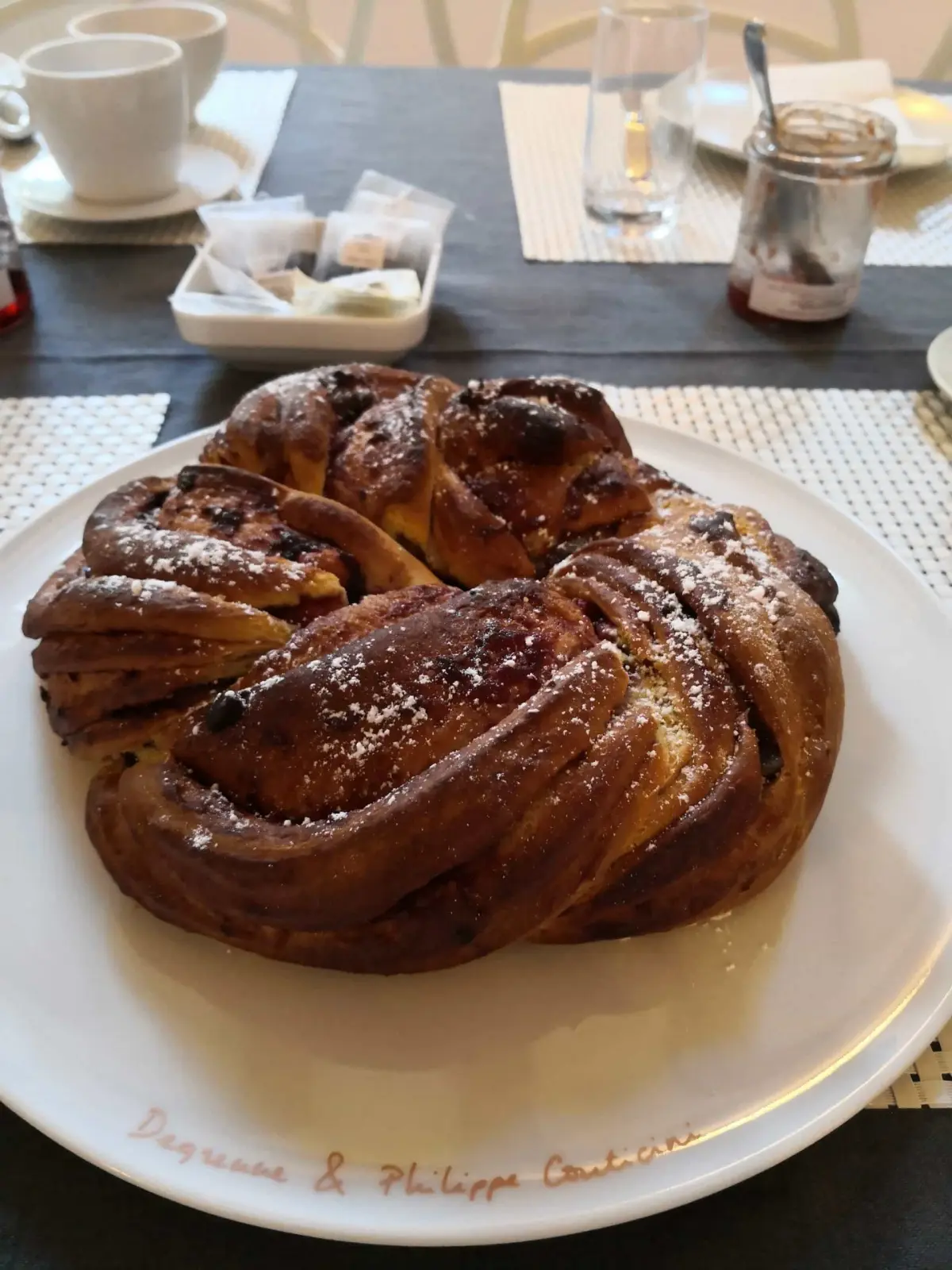 Braided brioche dusted with icing sugar on a white plate, cups and jam at breakfast, La Vie Voyage