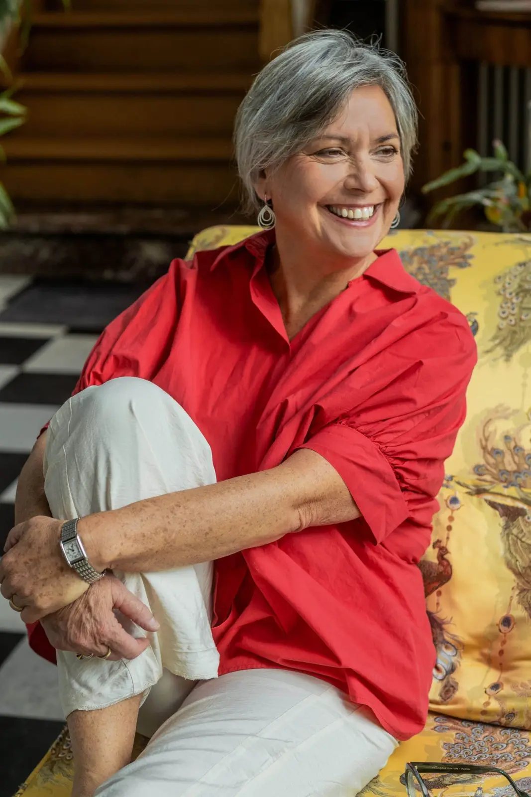 Muriel, host of La Villa Balat, smiling in a red shirt seated on a floral armchair
