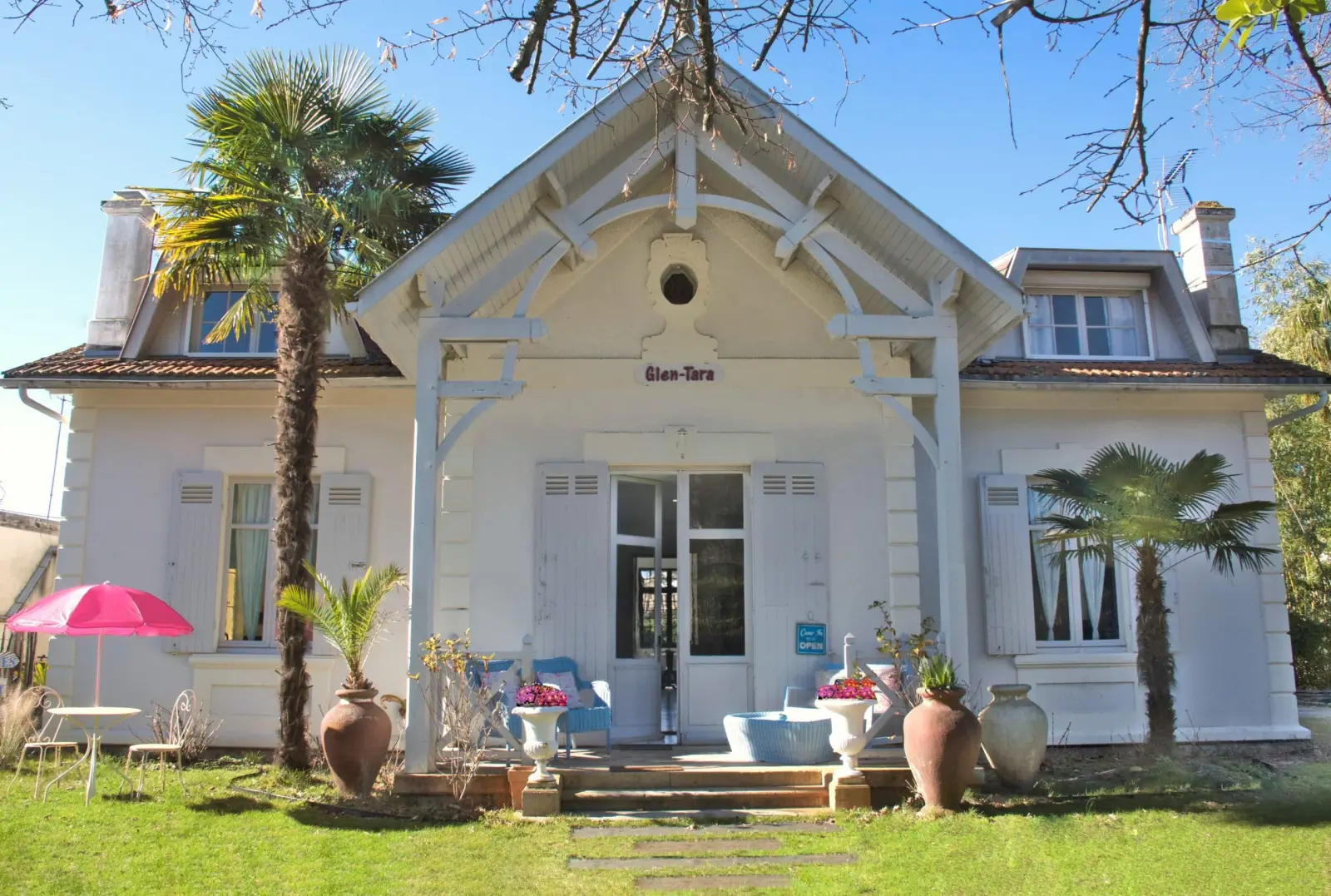 La villa Glen-Tara, façade blanche à boiseries, palmiers, jarres et parasol rose sur la pelouse